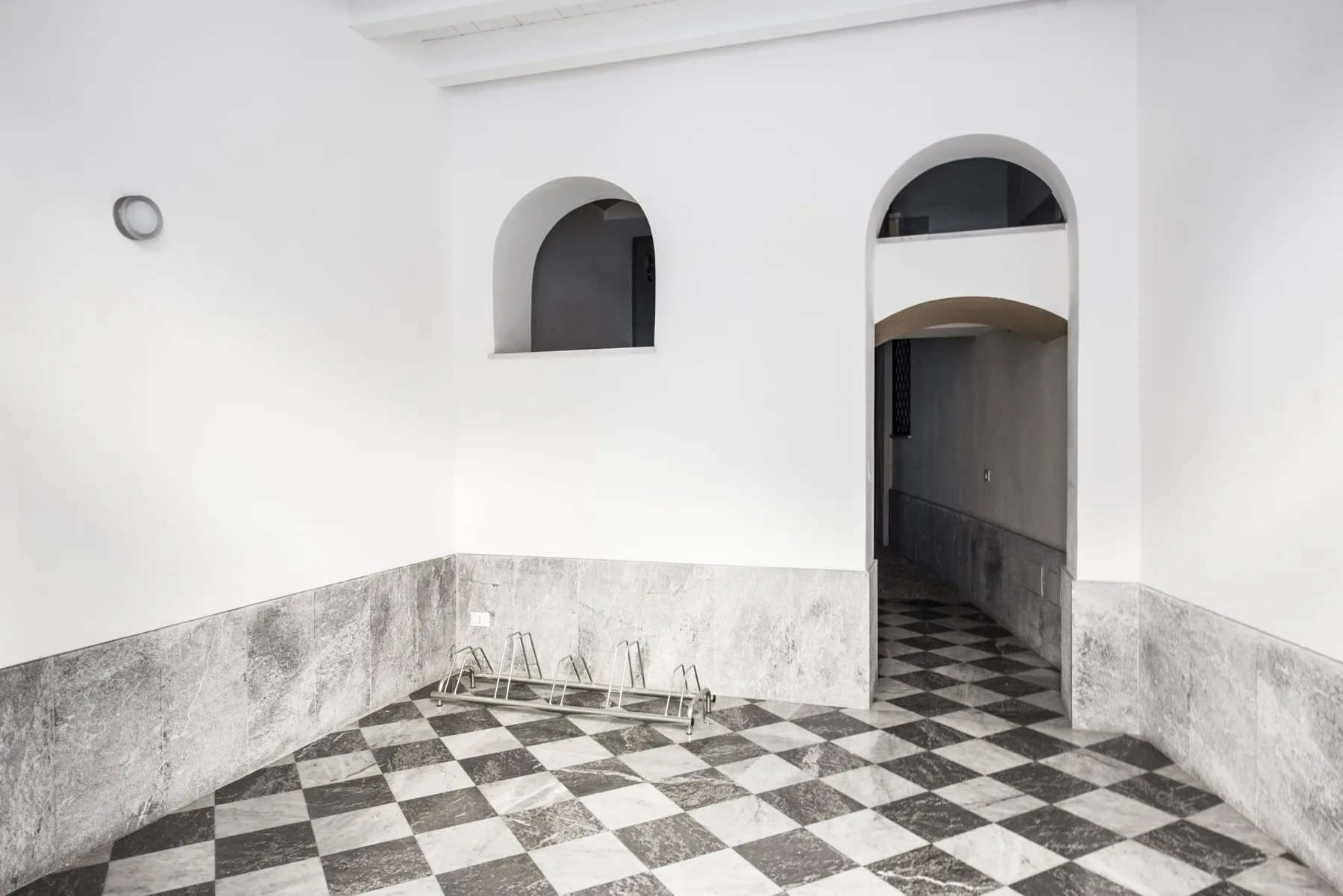Un pasillo interior con suelo de baldosas, arcos y una pequeña luz en la pared.