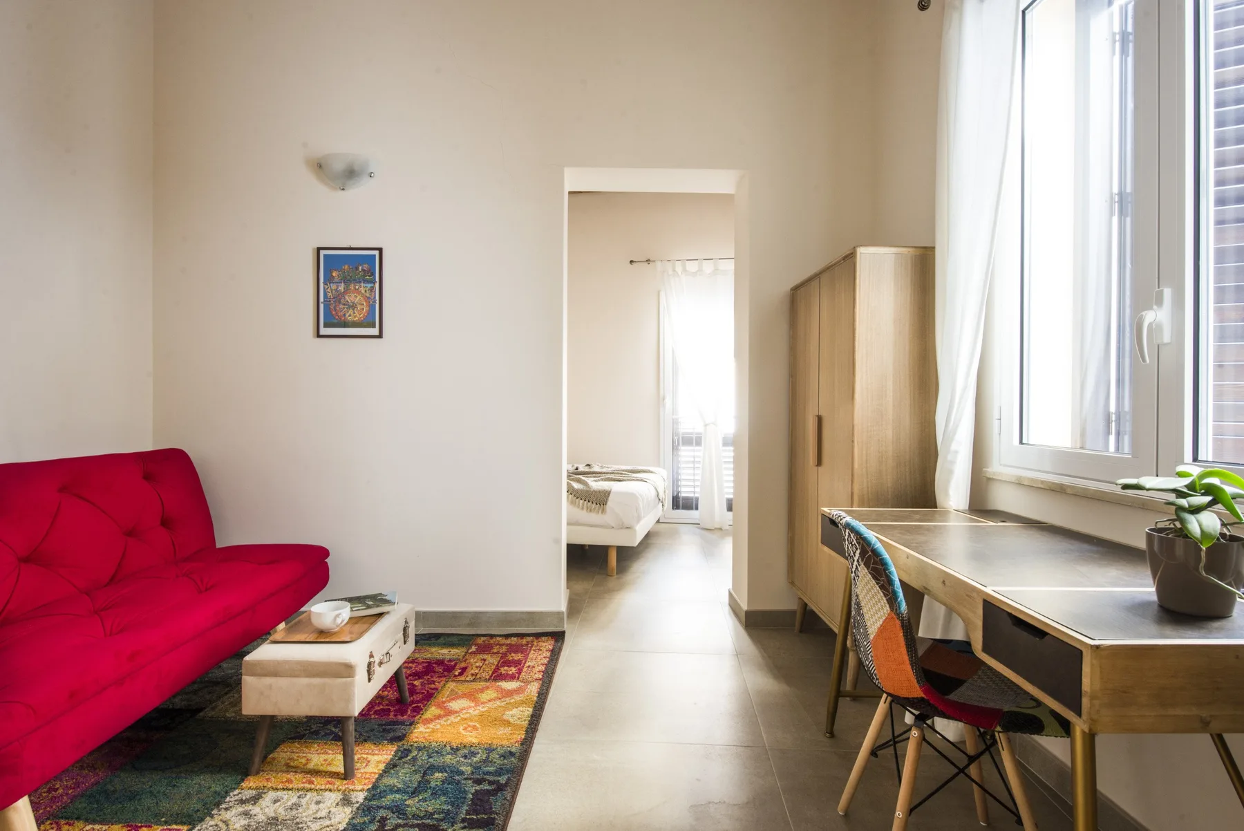 Una habitación luminosa con asiento rojo, un escritorio junto a la ventana y una puerta hacia un dormitorio.