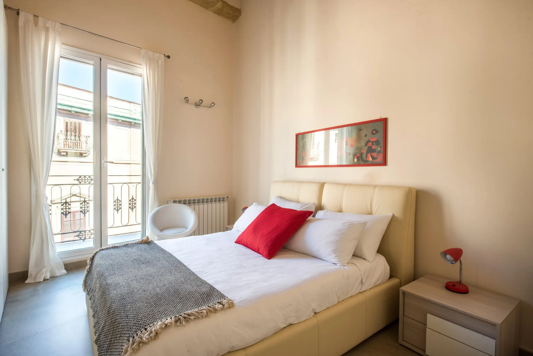 Un dormitorio con una cama, un cojín rojo y ventanas altas.
