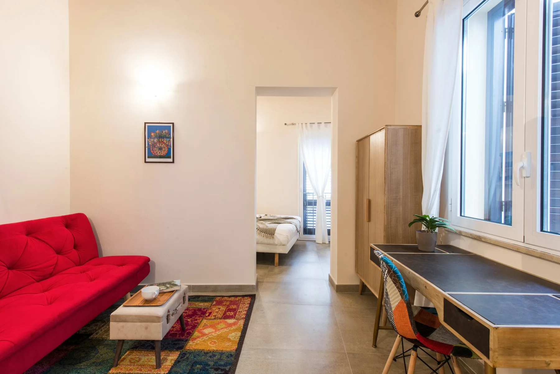 Una habitación luminosa con asientos rojos, un escritorio junto a la ventana y una puerta abierta al dormitorio.