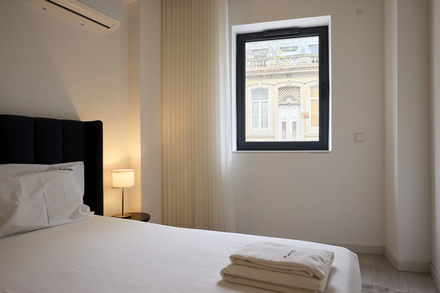 A bedroom with a neatly made bed and a window overlooking a neighboring building.