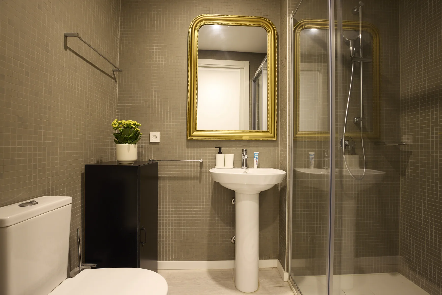 A bathroom with a pedestal sink, gold-framed mirror, and glass shower.