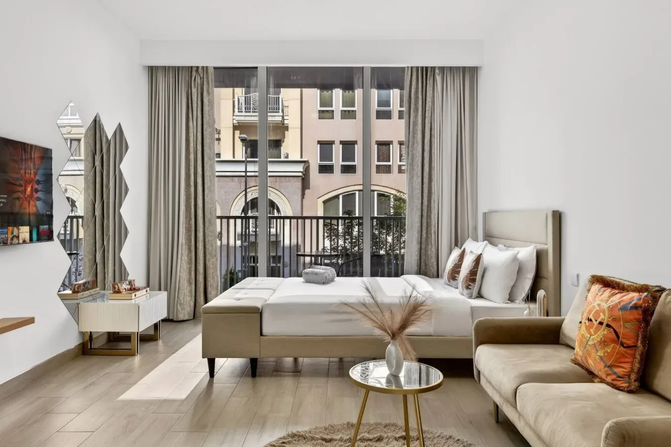 A bright living room with a bed, seating, and tall windows opening onto a balcony.