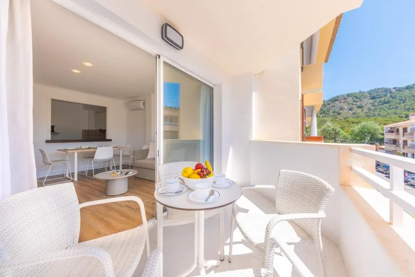A bright balcony with white seating and a small table overlooks a green hillside.