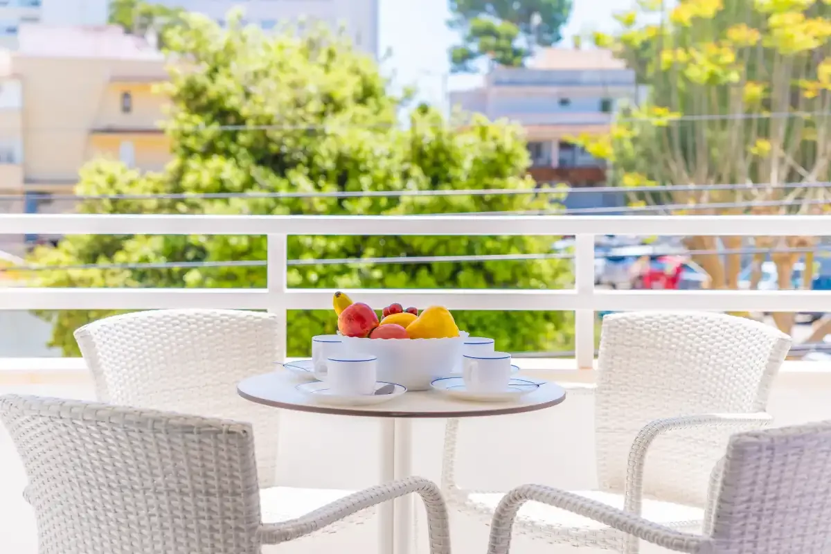 A balcony seating area with a small table and fruit bowl overlooks trees and nearby buildings.