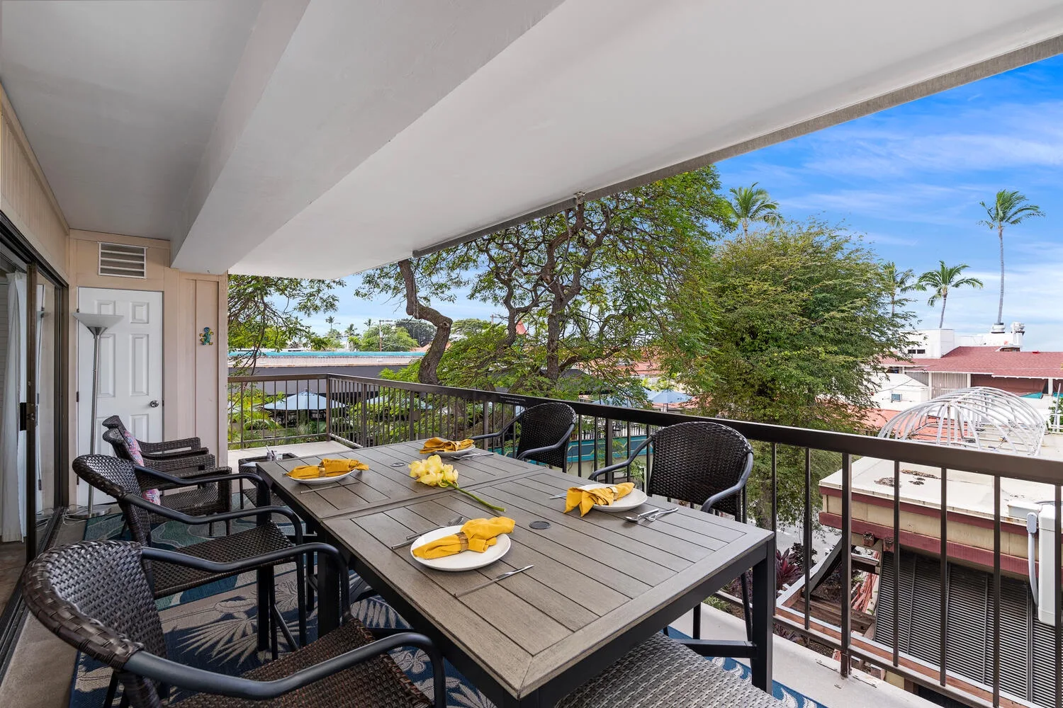 Un balcón cubierto tiene una mesa de comedor y asientos con vistas a árboles y edificios cercanos.