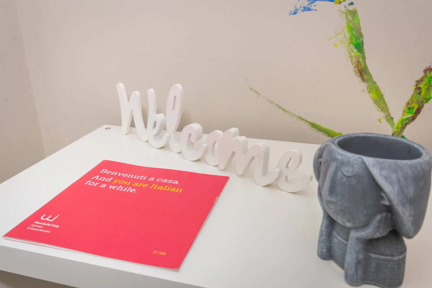 Una mesa blanca muestra un folleto rojo y un letrero decorativo de bienvenida junto a una figura de elefante gris.