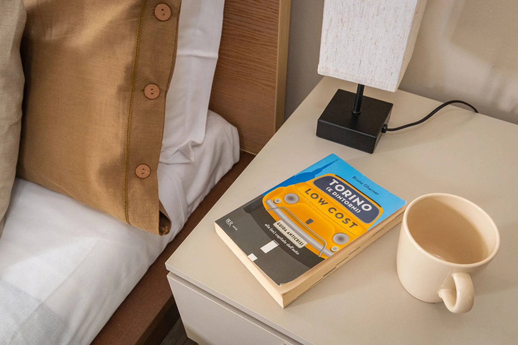 Una mesita de noche tiene una lámpara, un libro y una taza junto a la cama.