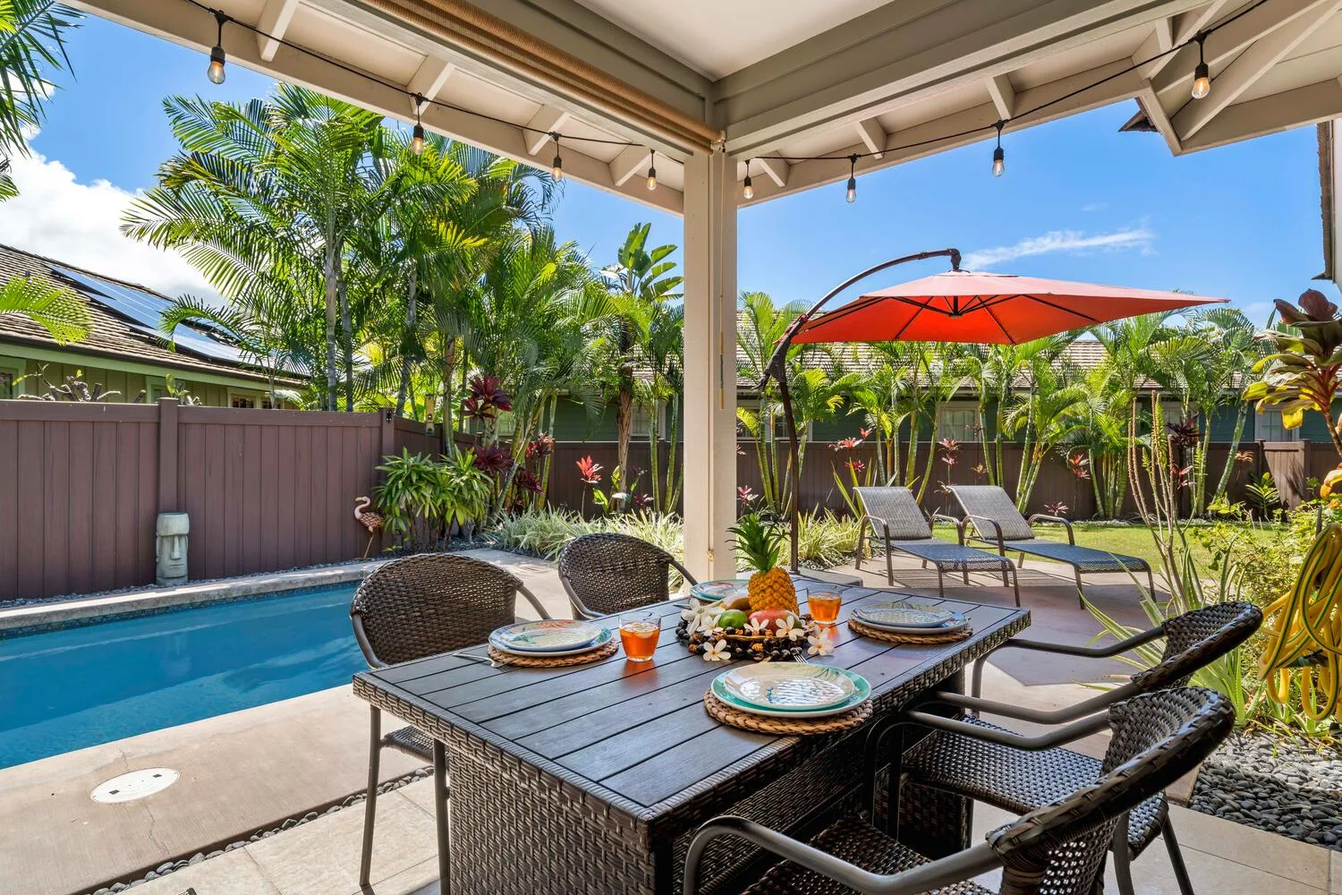A covered patio with a dining table overlooks a swimming pool and garden.