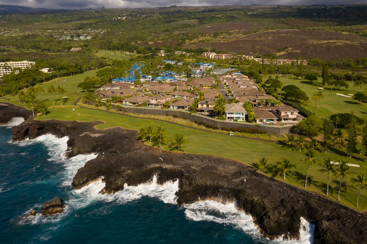 Aerial view of a coastal resort complex beside green grounds and rocky shoreline.