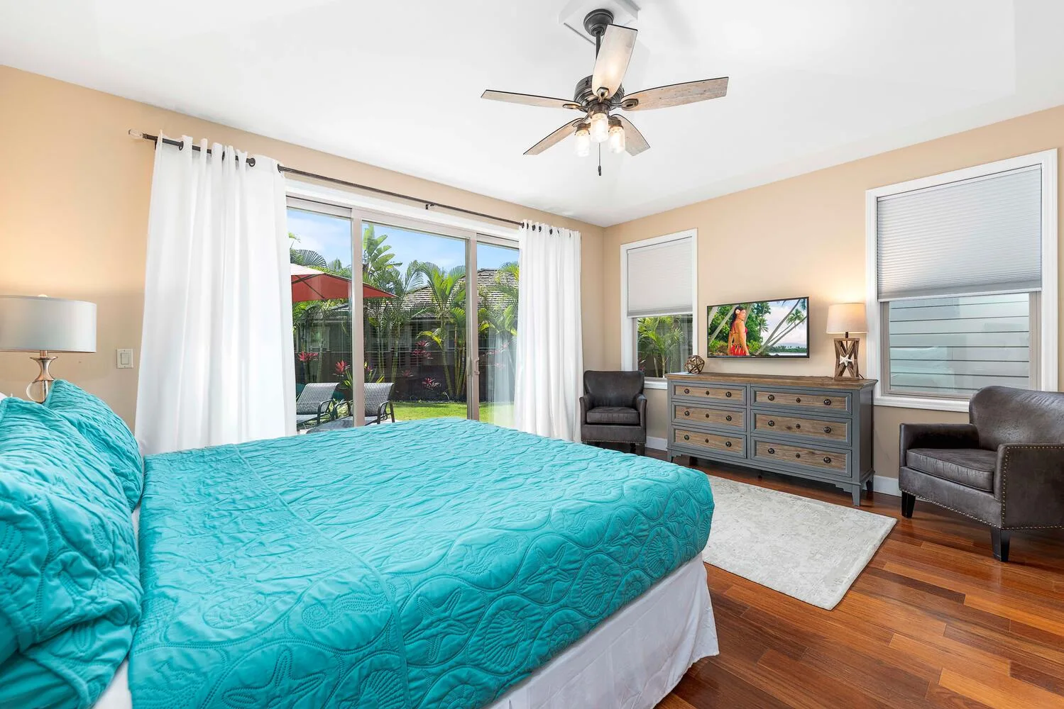 A bedroom with a turquoise bedspread, dresser, and windows overlooking greenery.