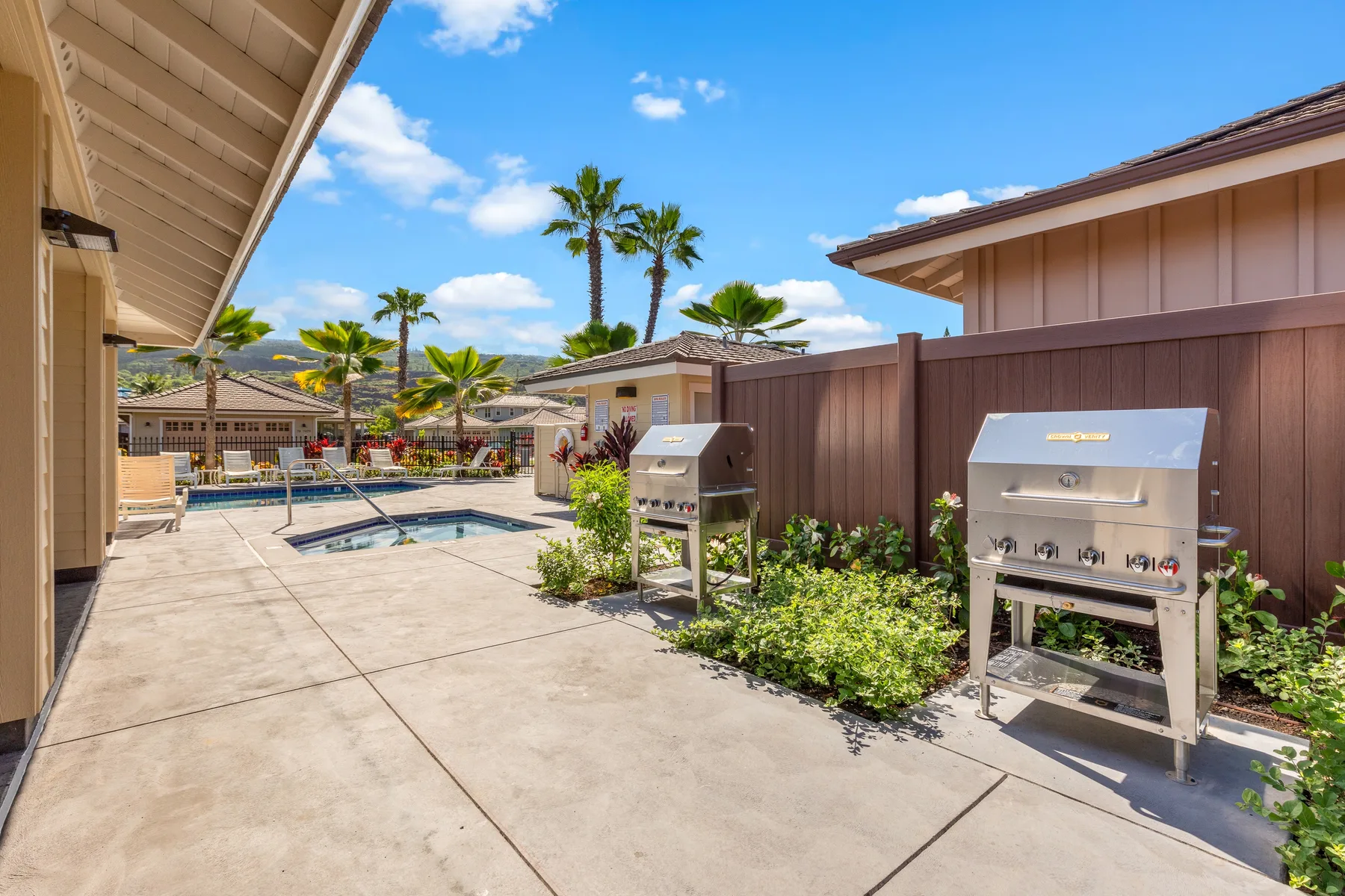A sunny outdoor patio has grills beside a swimming pool and palm trees.