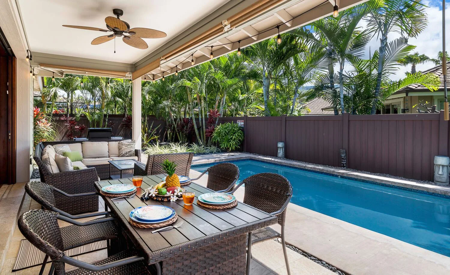 A covered patio with outdoor seating and a swimming pool beside tropical plants.