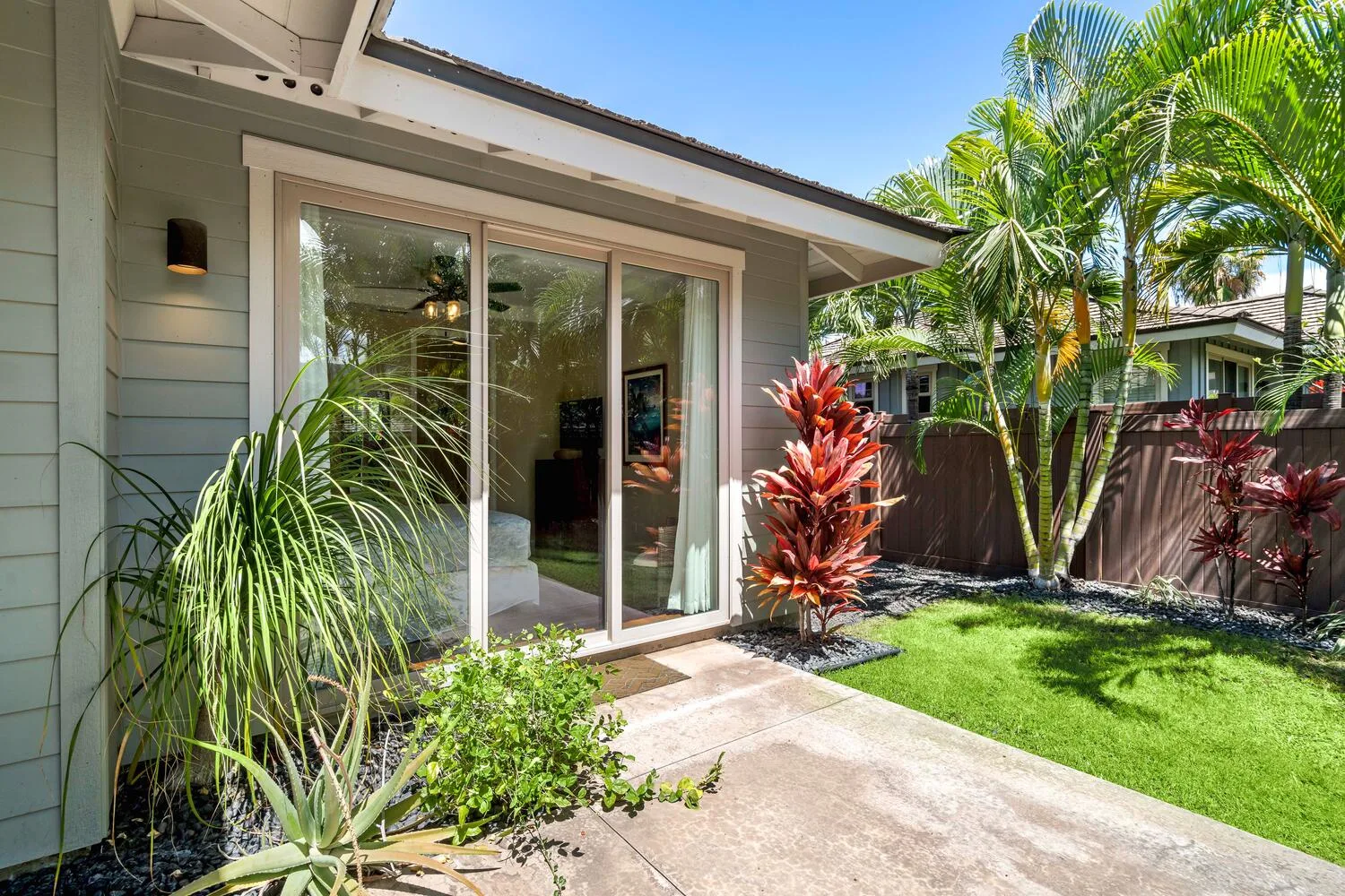 A sunny garden patio beside a house, with sliding glass doors and tropical plants.