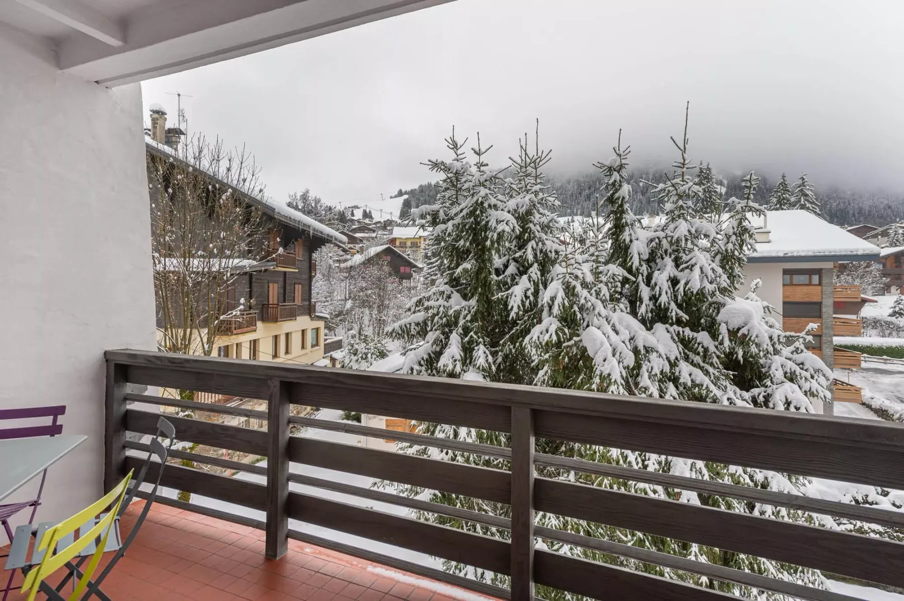 Un balcón da a árboles y edificios cubiertos de nieve en un entorno montañoso.