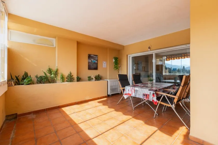 A covered terrace has a dining table and chairs beside sliding glass doors.
