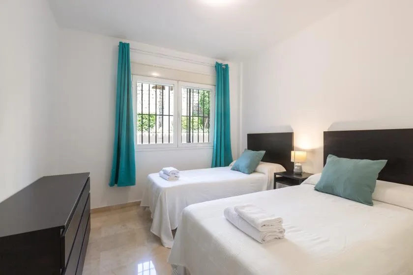 A bright bedroom with two beds, turquoise curtains, and a barred window.