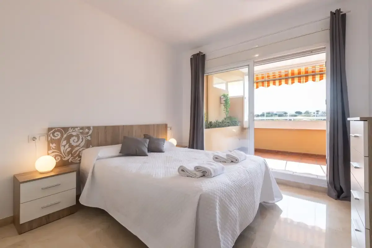 A bright bedroom with a neatly made bed beside an open balcony.