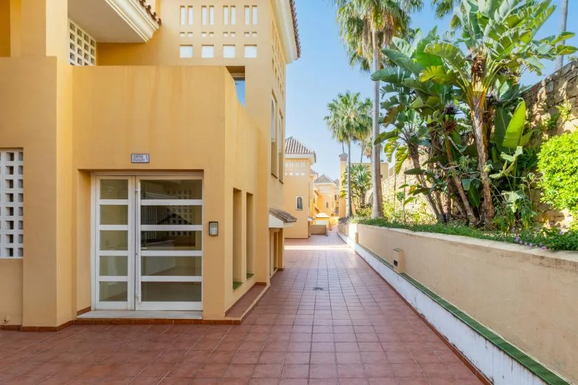 A tiled outdoor walkway runs beside a tan building and tropical plants.