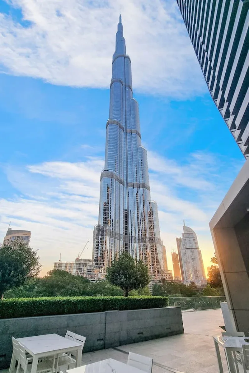 A tall glass skyscraper rises above a landscaped plaza under a blue sky.