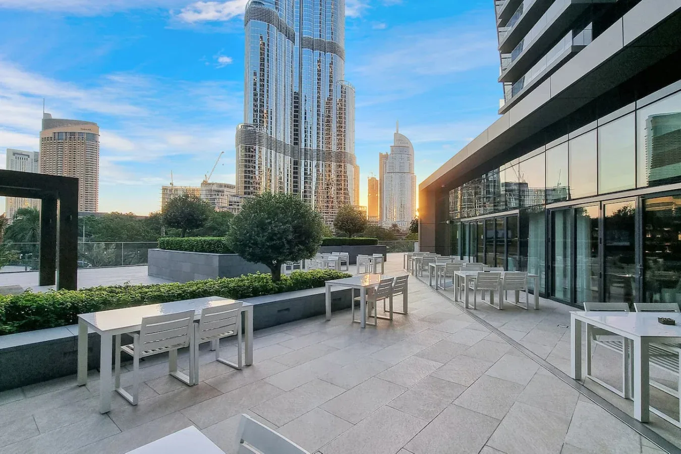 An outdoor terrace with tables and seating overlooks landscaped grounds and city towers.