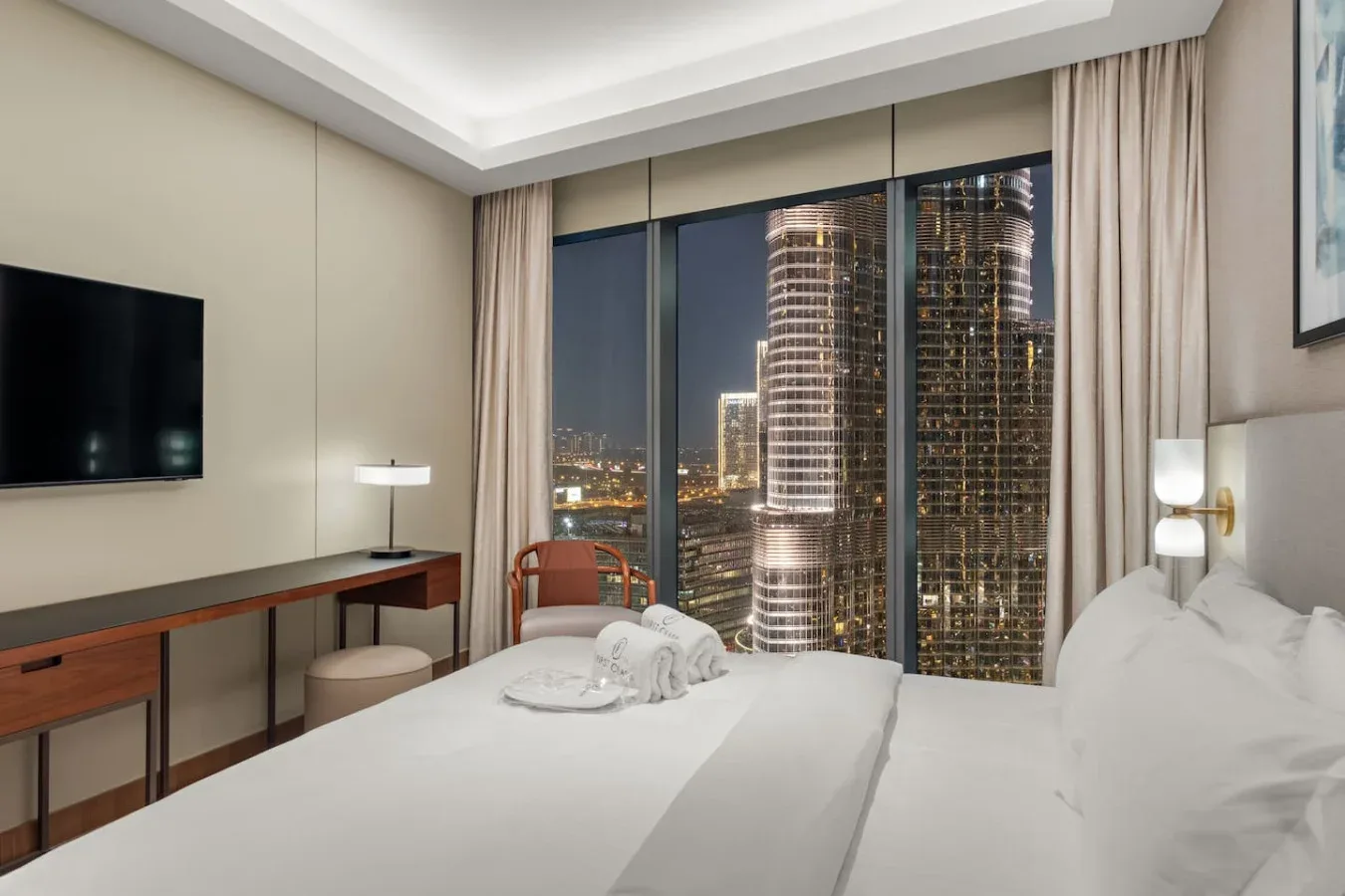 A bedroom with white bedding, a desk, and large windows overlooking city buildings.