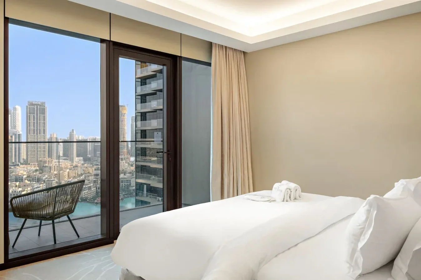 A bedroom with a neatly made bed beside floor-to-ceiling windows overlooking the city.