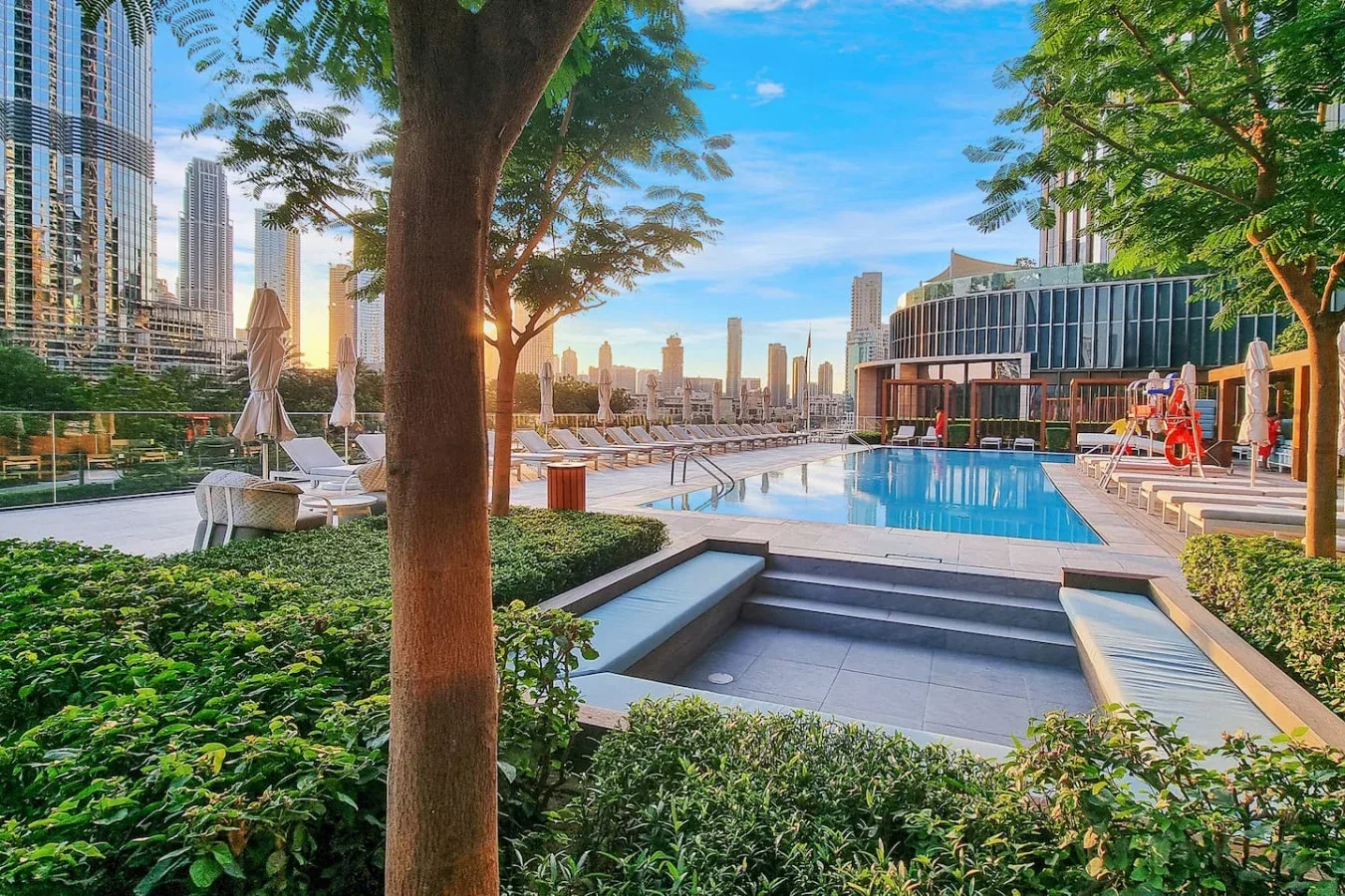 A landscaped outdoor pool with lounge seating and city buildings in the background.