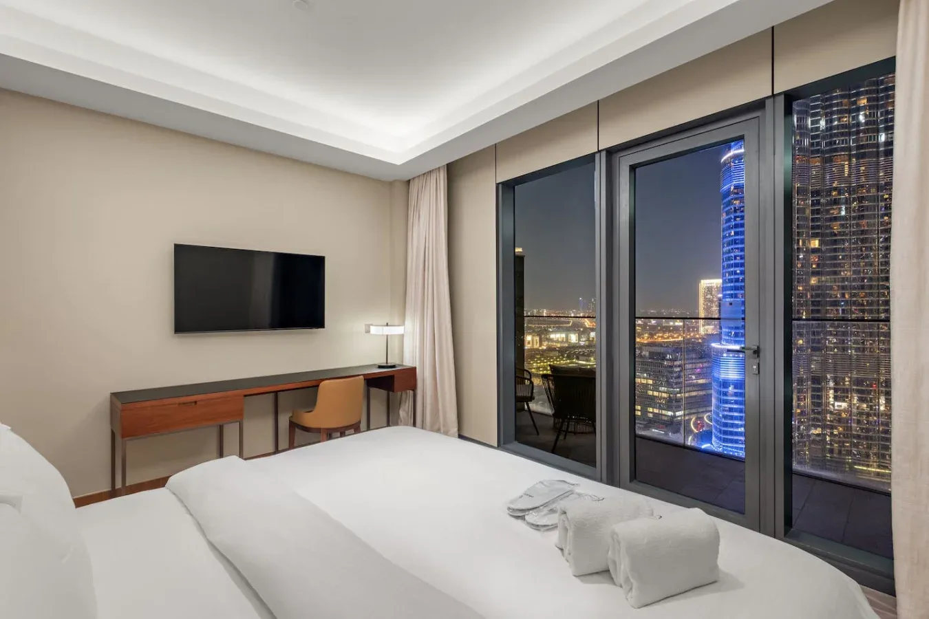 A modern bedroom with a bed, desk, and floor-to-ceiling windows overlooking city lights.