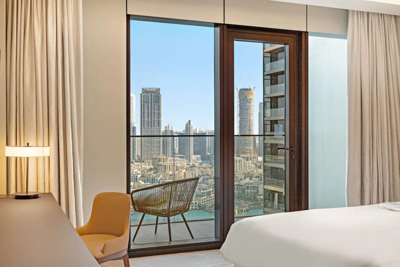A bedroom with a bed beside floor-to-ceiling windows overlooking city buildings.