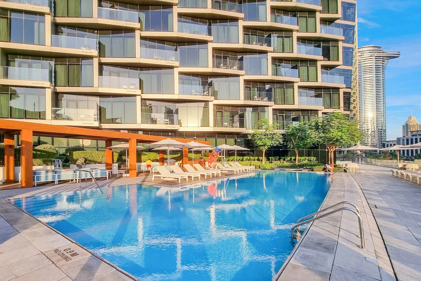 An outdoor swimming pool beside a modern apartment building, with lounge seating along the deck.