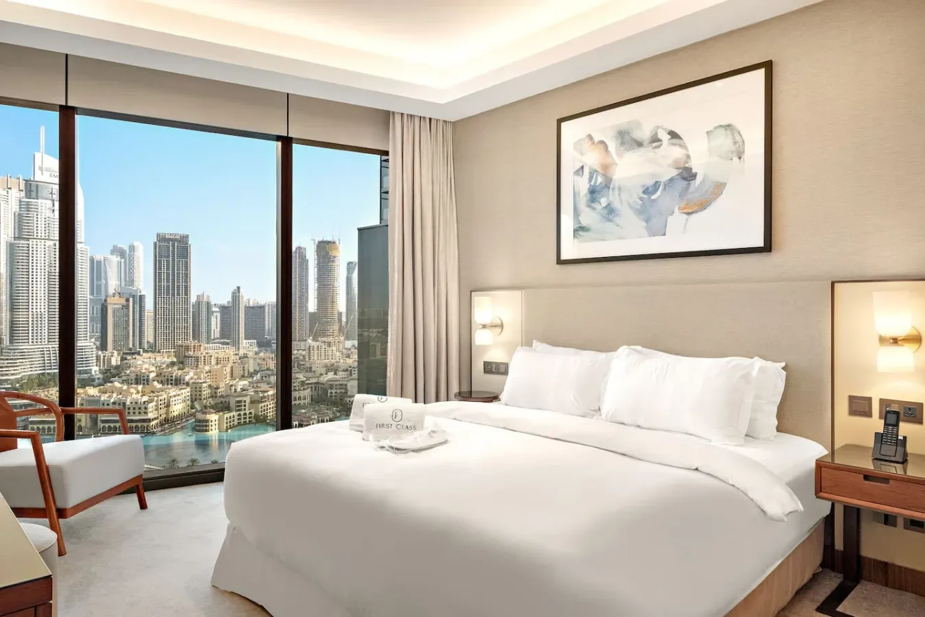 A bedroom with a large bed beside floor-to-ceiling windows overlooking city buildings.