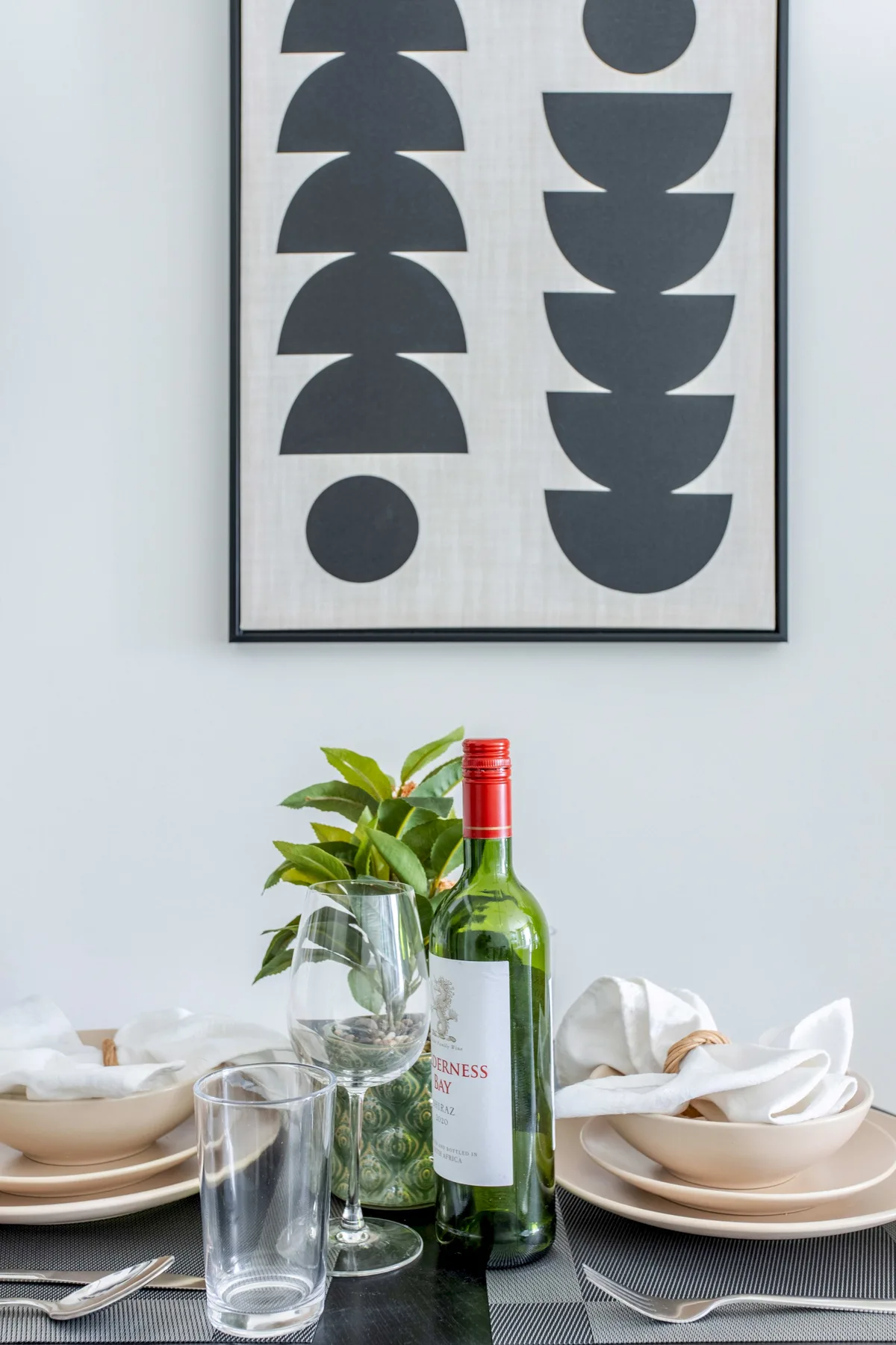 Una mesa de comedor está puesta con platos, vasos, servilletas y una planta frondosa bajo un cuadro abstracto.