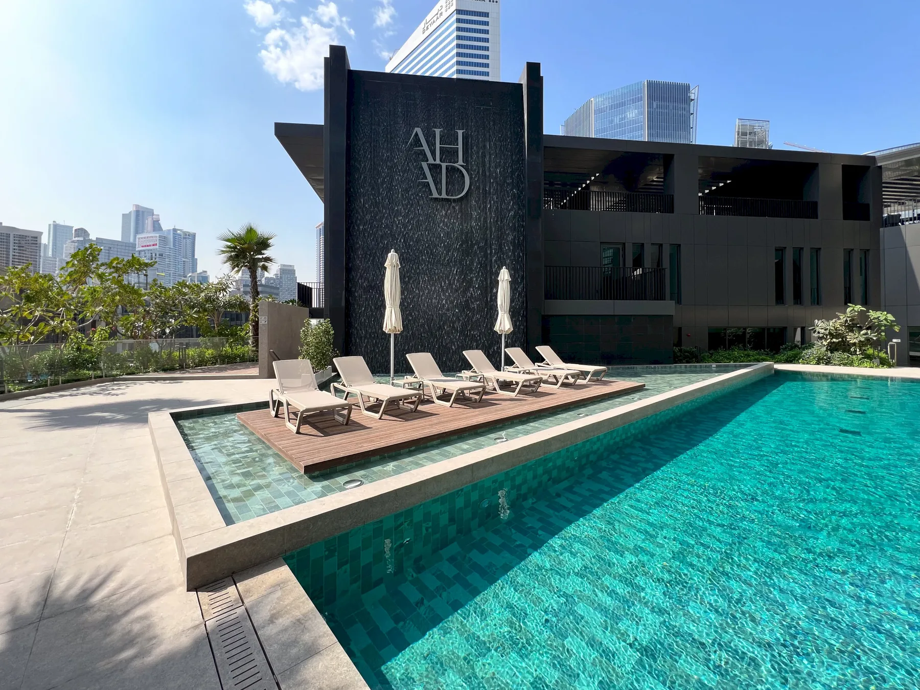 Una piscina exterior junto a un edificio moderno oscuro, con tumbonas a lo largo del borde.