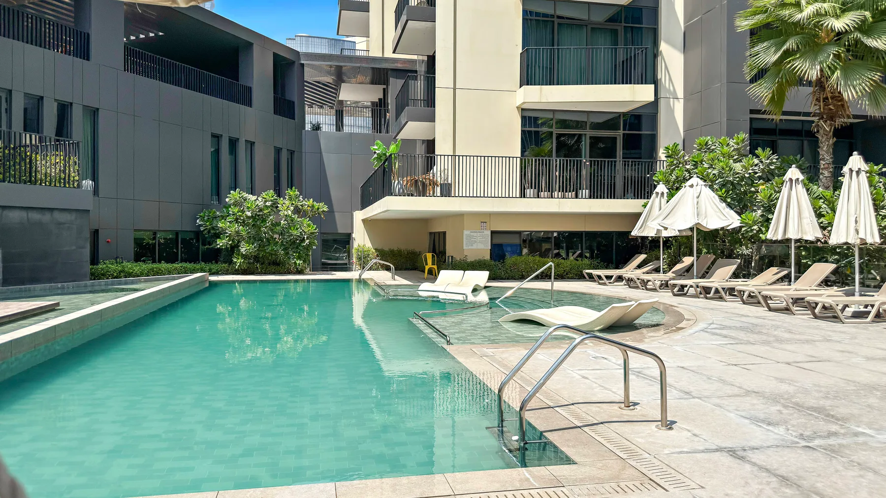 Una piscina al aire libre junto a una terraza pavimentada con tumbonas y sombrillas.