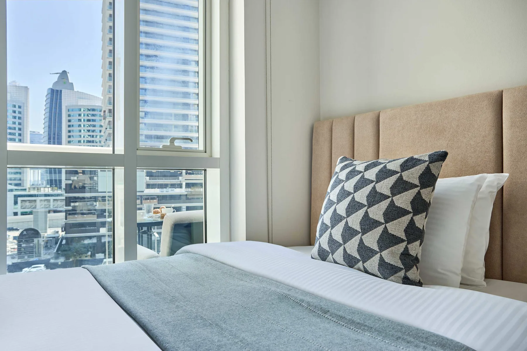Un dormitorio con una cama junto a grandes ventanas con vistas a edificios cercanos.