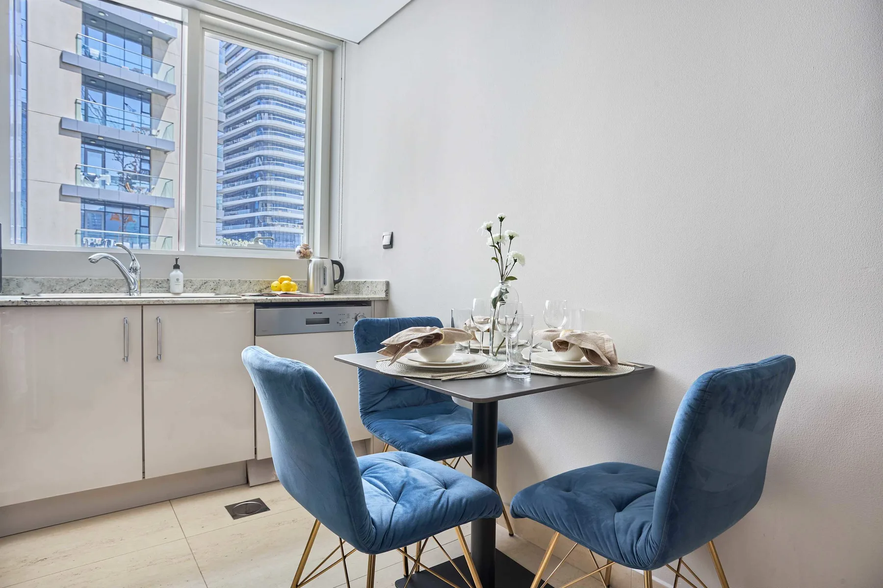 Comedor con una mesa puesta junto a grandes ventanas con vistas a edificios vecinos.