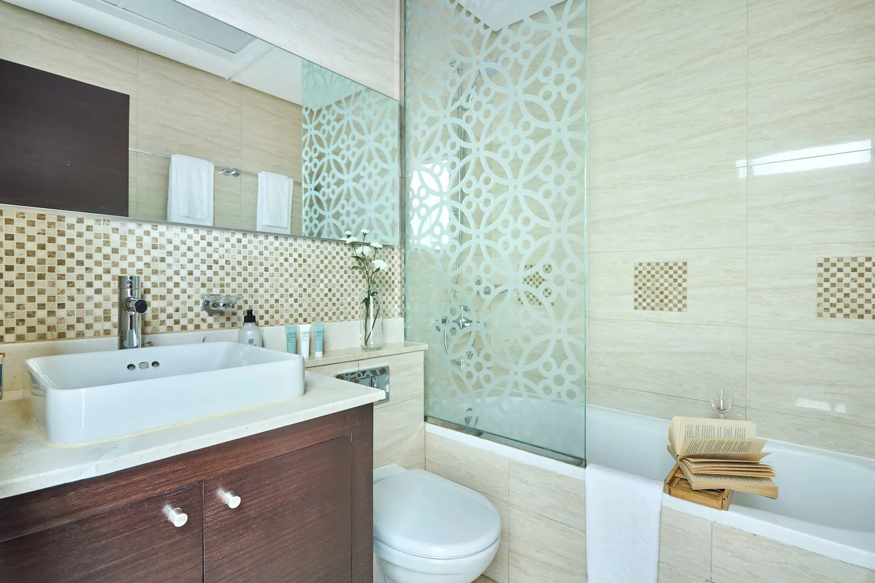 Un baño luminoso con lavabo, inodoro y bañera tras un panel de vidrio decorado.
