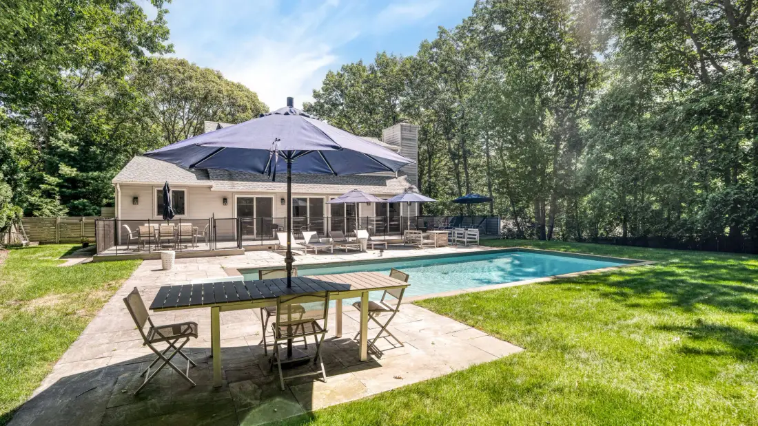 Una piscina en el patio trasero está junto a un patio con asientos y una sombrilla.