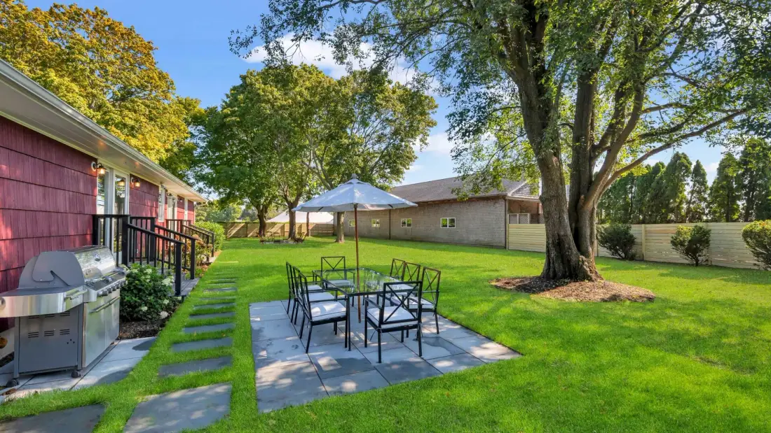 A backyard patio has outdoor seating beneath a large tree, with a grill beside the house.