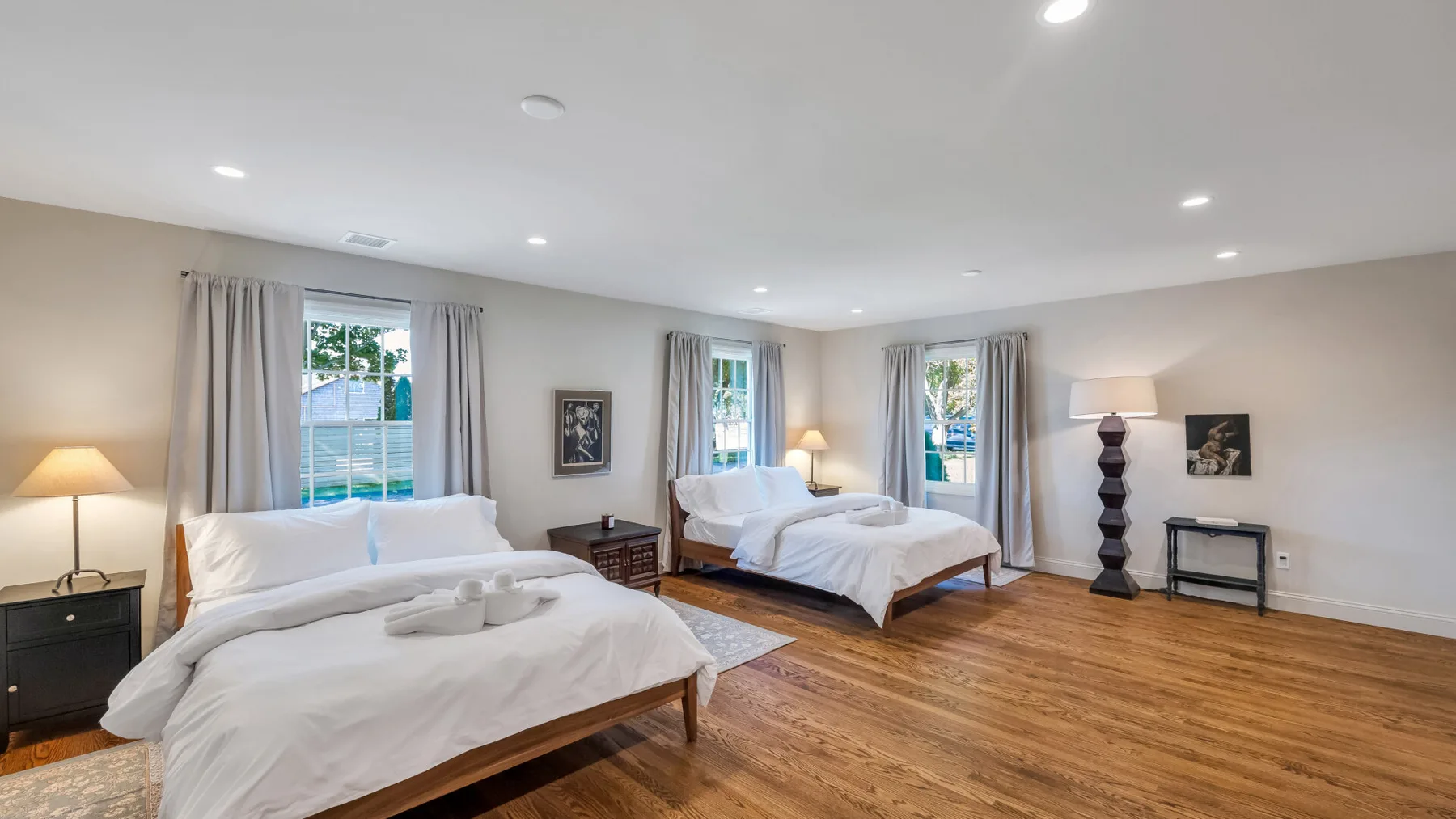 A bright bedroom with two beds, curtained windows, and wood flooring.