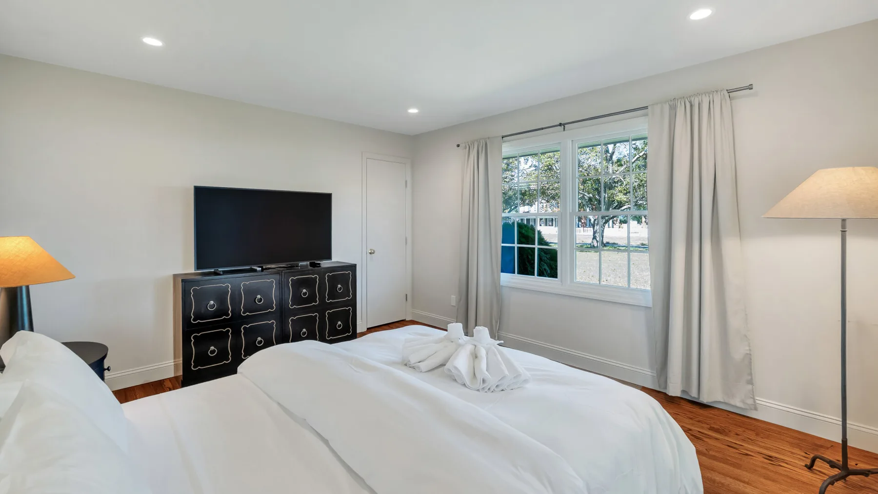 A bright bedroom with white bedding, a dresser, and a curtained window.
