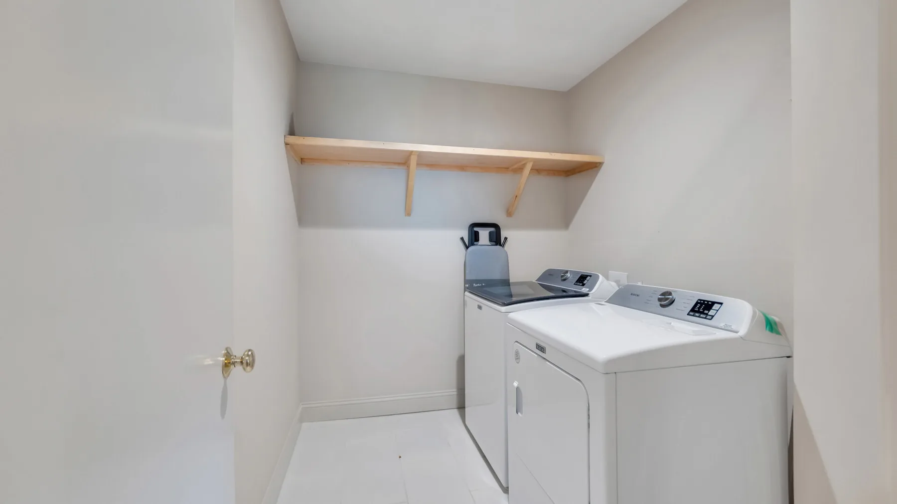 A laundry room with a washer and dryer beneath a wooden shelf.
