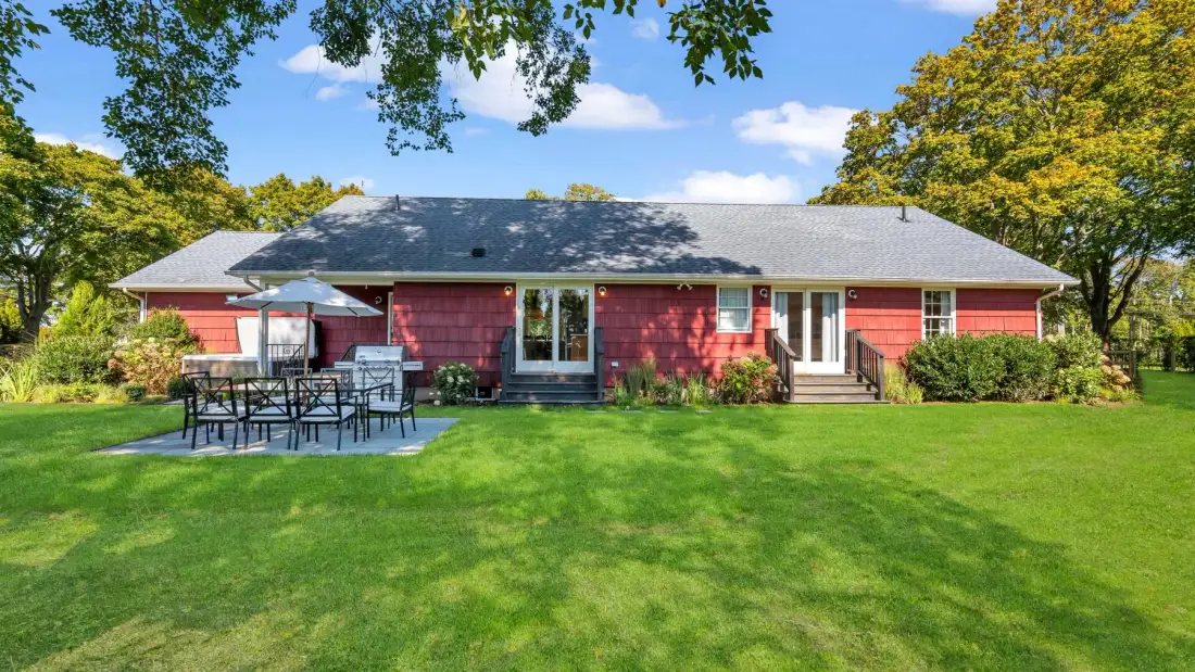 A red house stands beyond a broad green lawn, with outdoor seating nearby.