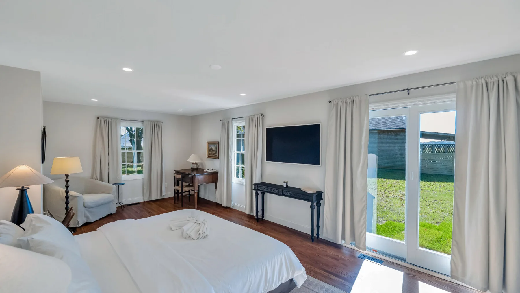 A bright bedroom with a large bed, a television, and glass doors opening onto a lawn.