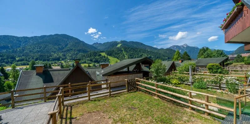 A fenced garden overlooks wooden houses and green mountains beneath a blue sky.