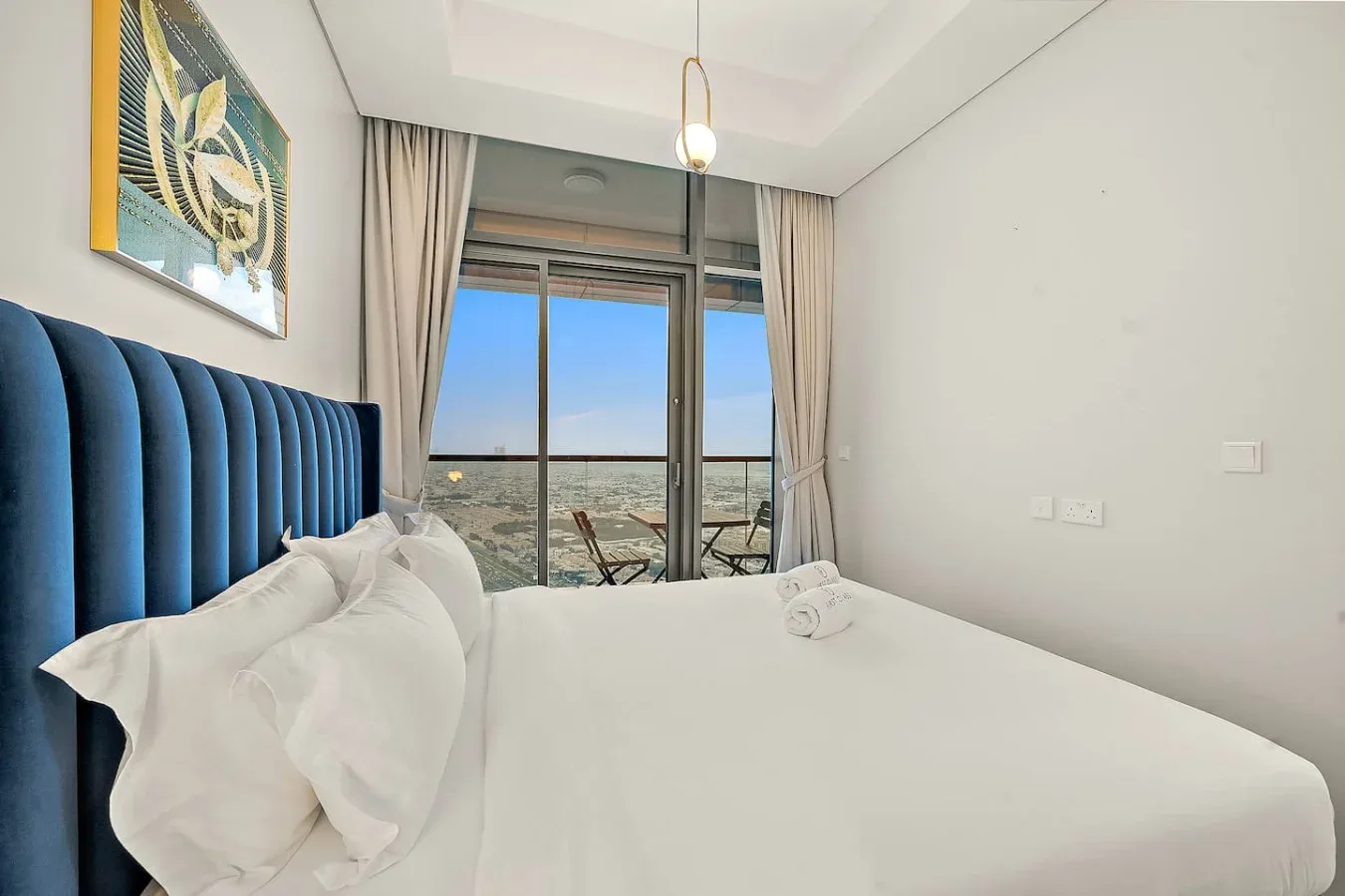 Un dormitorio luminoso con cama blanca, cabecero azul tapizado y puertas de vidrio.