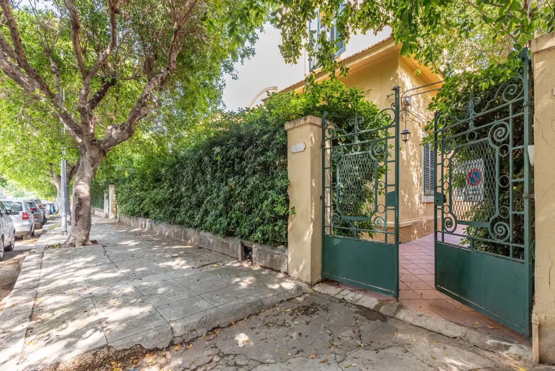 A leafy sidewalk runs beside a tall hedge and an open decorative gate.