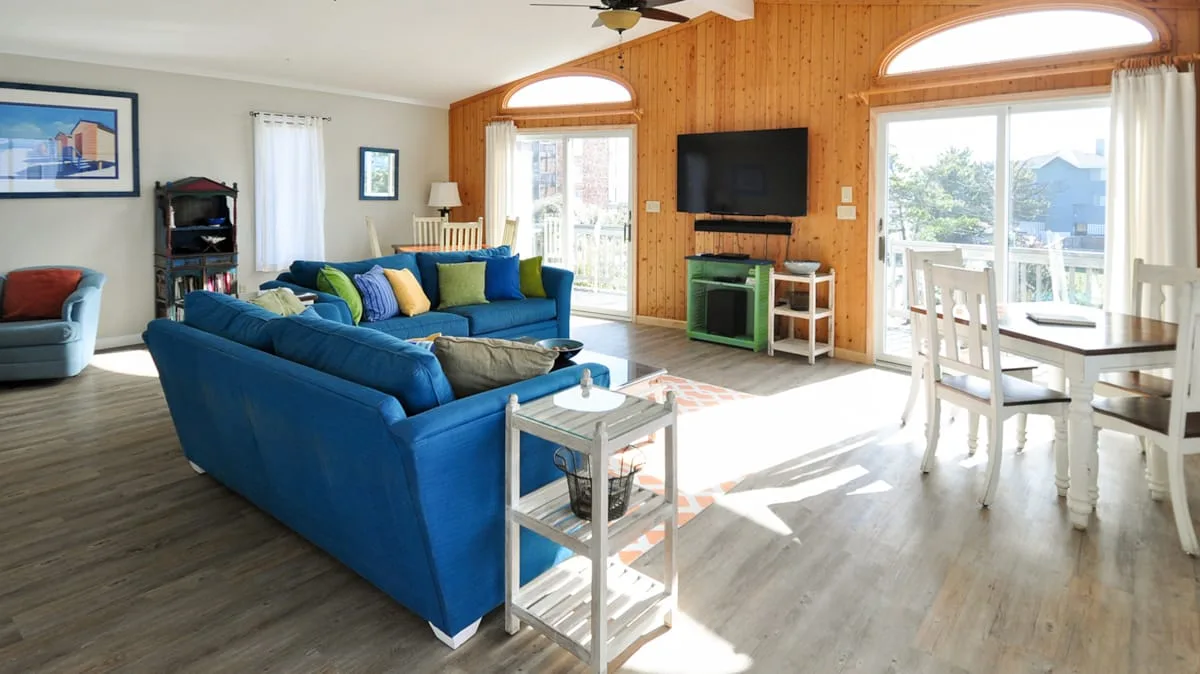 Una sala luminosa tiene asientos azules, un televisor y puertas que dan a un patio.
