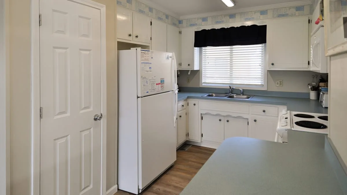 Una cocina con gabinetes blancos, un refrigerador y una encimera bajo la ventana.